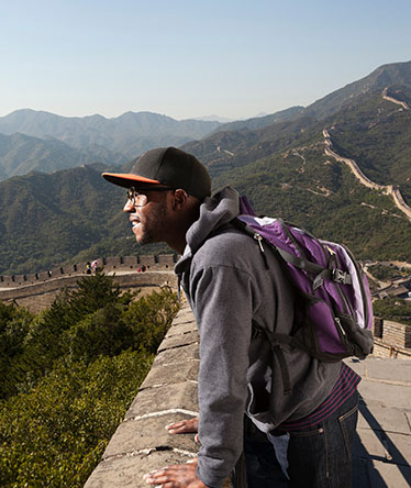 Man on the great wall of China, wearing glasses, cap, grey sweater and purple backpack.